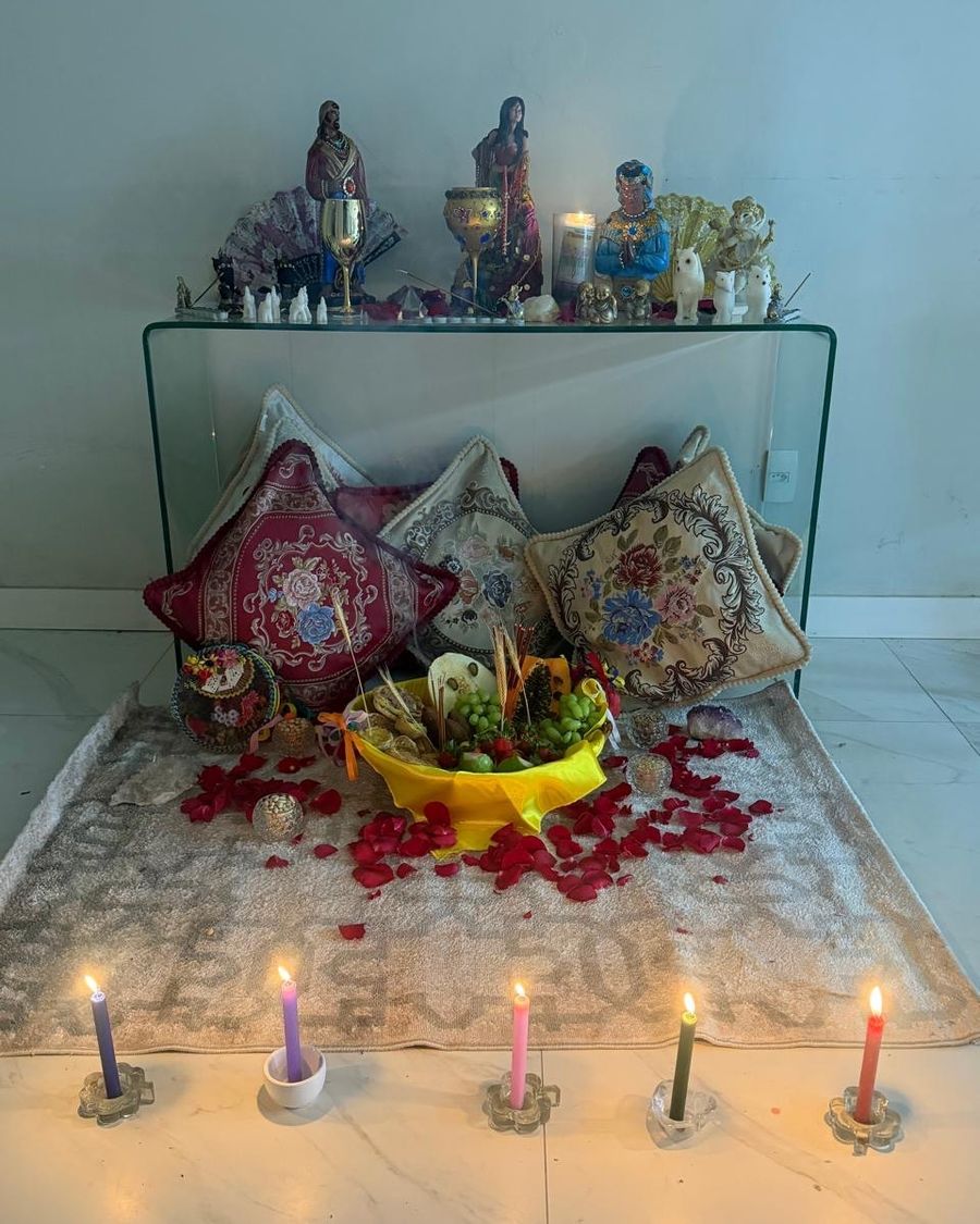 Altar com imagens de ciganas, almofadas bordadas, cesta de frutas, pétalas de rosa e cinco velas coloridas acesas