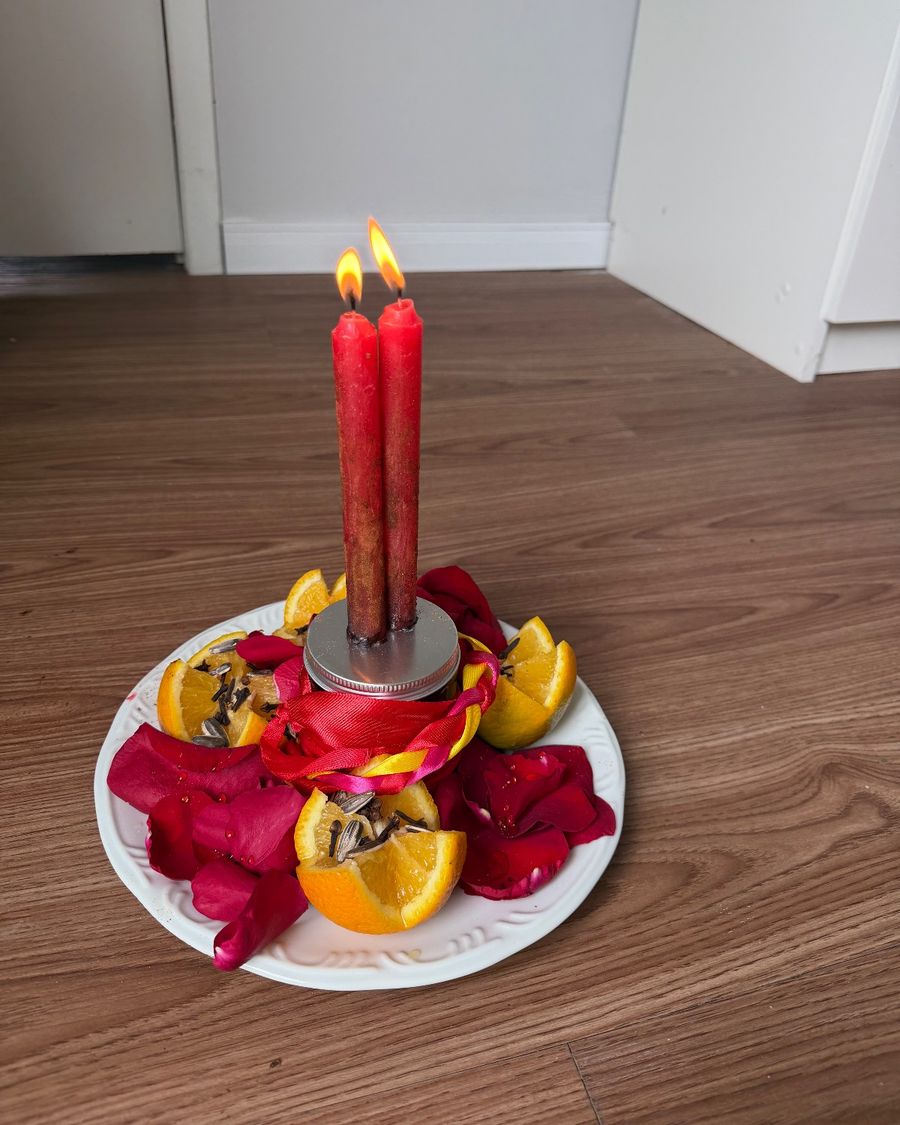 Magia de Hoodoo: velas vermelhas acesas sobre pote amarrado com fitas, laranjas com cravo e pétalas de rosa