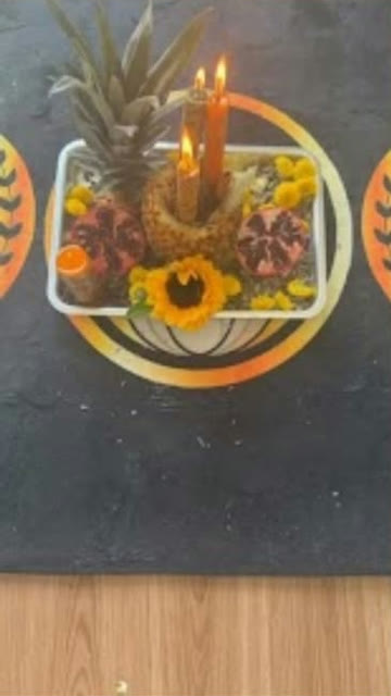 Oferenda com abacaxi, velas acesas, romãs, girassol e flores amarelas sobre um tapete com sol dourado